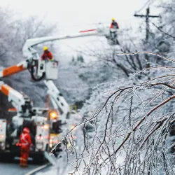 Utility workers in bucket trucks repairing ice-covered power lines on a snowy winter road with frozen tree branches.