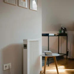 White portable heater with digital control on wooden floor next to small table and wall art in cozy room.