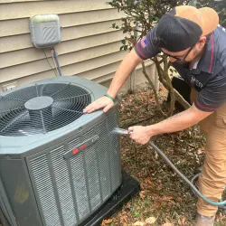 Technician cleaning an outdoor air conditioning unit with a hose beside a house exterior and bushes.