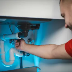 Plumber fixing white pipes under a sink with blue lighting highlighting the plumbing system.