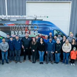 Group photo of Dayco Systems employees standing outside with company vans near industrial building.