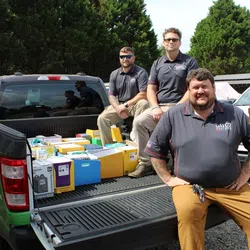 Three men in matching Dayco Systems shirts sit on the back of a pickup truck filled with organized office supplies outdoors.
