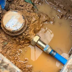 Water meter and blue pipe connection partially submerged in muddy water in a dirt excavation site.