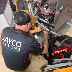 Technician in Dayco Systems shirt repairing HVAC unit with Milwaukee tool bag nearby on a concrete floor.