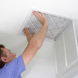 Man in blue shirt replacing or inspecting a ceiling air filter behind an open vent cover in a white room.