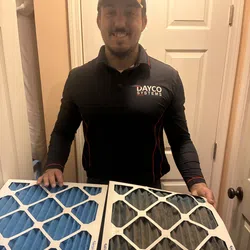 Man in Dayco Systems uniform holding new blue and used dirty HVAC air filters side by side indoors.