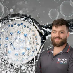 Plumber in Dayco Systems shirt stands smiling in front of close-up showerhead with water splashing and bubbles.