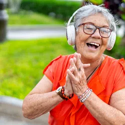 Joyful elderly woman in orange shirt wearing headphones and clapping hands outdoors in a garden.