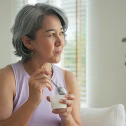 Mature woman with gray hair eating yogurt on a couch in a bright, cozy living room with plants.