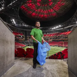 Worker in green shirt holding a blue trash bag inside a large, empty football stadium with red seats and illuminated field.
