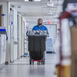 Janitor in blue uniform pushing a large black trash bin down a hospital corridor with medical equipment.