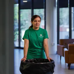 Woman in green shirt holding a trash bag in a modern, well-lit living room with large windows and brown chairs.