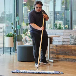 Man wearing glasses mopping wooden floor in modern indoor cafe with large windows and cozy seating area.