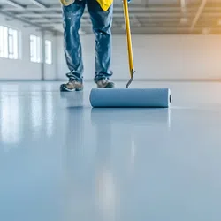 Worker applying blue paint smooth finish on large empty indoor floor with paint roller