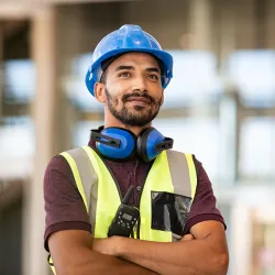 Confident construction worker in hard hat and safety gear, standing with arms crossed indoors.