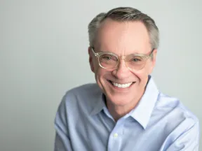 Smiling middle-aged man with glasses wearing a light blue shirt against a plain gray background.