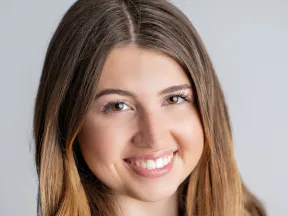 Close-up of a smiling young woman with long light brown hair and clear skin against a gray background