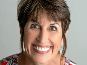 Smiling middle-aged woman with short brown hair and bright eyes wearing colorful patterned clothing.