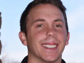Young man smiling outdoors with brick wall and clear sky in the background.
