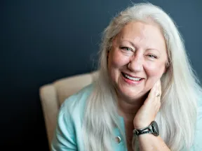 Smiling elderly woman with long white hair in blue top sitting in chair against dark background