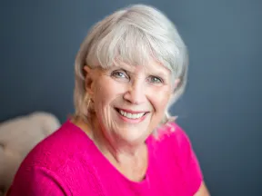 Smiling elderly woman with white hair wearing a bright pink top against a blue background.