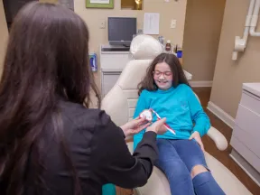 Dentist demonstrating proper tooth brushing technique to smiling girl sitting in dental chair at clinic.
