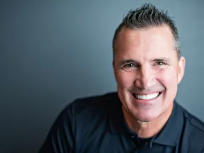 Smiling middle-aged man with short hair wearing a black polo shirt against a gray background.