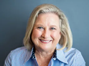 Smiling mature woman with blonde hair wearing a blue striped shirt against gray background.