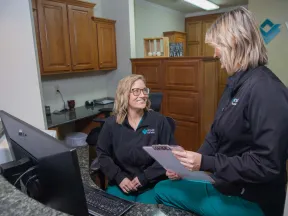 Two female medical professionals in black jackets discussing patient files in a modern clinic office.