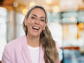 Smiling woman with long hair in pink blazer and headband in a bright, blurred indoor setting