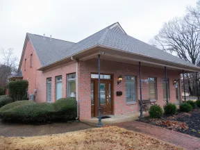 Single-story red brick building with peaked roof and large wooden door surrounded by bushes and a paved walkway.
