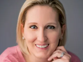 Close-up of smiling blonde woman with blue eyes wearing a pink top and a ring on her hand against gray background