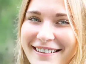 Close-up of a smiling young woman with blonde hair and blue eyes against a blurred green background