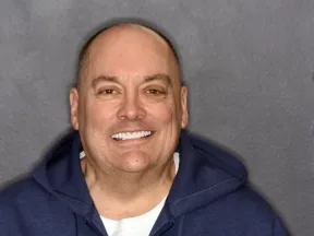 Smiling bald man wearing a navy blue hoodie against a gray wall background.