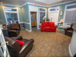 Cozy waiting room with red sofa, brown leather chairs, potted plants, lamps, and large windows with blinds.
