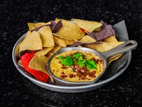 Plate of multicolored tortilla chips with a skillet of cheese dip garnished with cilantro on a dark surface