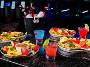 Plates of colorful tortilla chips with dips and mixed drinks on a dark speckled table in a dim bar setting.