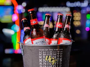 Bucket with UCF Knights and Corona branding holding six Budweiser beer bottles with colorful bokeh background.