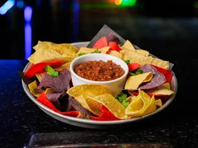 Plate of multicolored tortilla chips served with a bowl of chunky salsa dip on a dark surface.