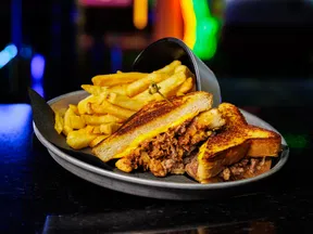 Grilled pulled pork sandwich with melted cheese served with golden French fries on a metal plate.