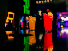 Two colorful cocktails with fruit garnishes on a bar counter with neon lights and reflections.