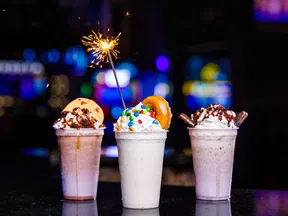 Three colorful milkshakes topped with whipped cream, candy, cookie, donut, and a lit sparkler on a dark background.