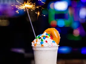 Creamy milkshake topped with whipped cream, colorful candy, a donut, and a lit sparkler on a black surface.