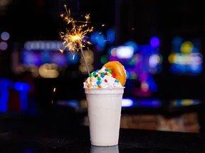 Creamy milkshake with whipped cream, colorful candy, donut, and lit sparkler in a dimly lit setting.