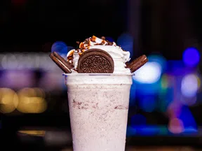 Creamy cookies and cream milkshake topped with whipped cream, chocolate drizzle, and Oreo cookies on black surface with lights.