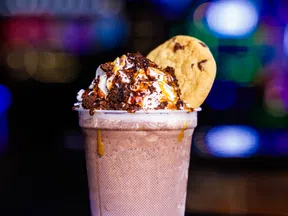 Chocolate milkshake topped with whipped cream, chocolate drizzle, cookie crumbs, and a chocolate chip cookie on the side.