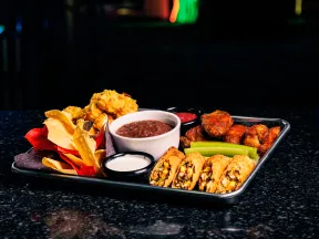 Tray with assorted appetizers including chips, dipping sauces, chicken wings, celery, and stuffed rolls on black surface.