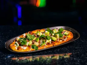 Flatbread topped with avocado, cilantro, ground meat, and sauce on a dark oval plate against a dark background