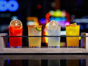 Four colorful cocktails with citrus garnishes served in a wooden tray against a blurred bar background.