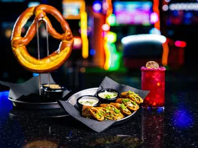 Soft pretzel on stand with dipping sauces, stuffed jalapeno poppers, and a red cocktail on a bar counter with colorful lights.